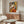 May include: Framed cubist-style artwork featuring a seated woman in geometric orange, yellow, and black shapes holding a fan, displayed above a walnut wood credenza with sliding doors. Modern living room setting with cream upholstered chair, glass oval coffee table, and white spherical table lamp. Minimalist decor with neutral walls and natural lighting.