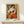 May include: Framed poster displaying a cubist-style woman in geometric orange, yellow, white and black shapes, holding a colorful angular fan. The artwork sits on a light wood shelf against a cream wall, with a small white wrapped gift box positioned in front. Natural lighting creates soft shadows on the minimalist setting.