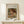May include: Framed art print of Pierre Auguste Renoir painting showing young child with reddish-brown hair reading a book, rendered in soft impressionist style with warm earth tones. Print displayed in light wood frame with beige matting on white wooden surface alongside small wrapped white gift box with evergreen sprig. Cream colored wall background with natural lighting.