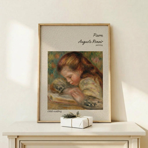 May include: Framed art print of Pierre Auguste Renoir painting showing young child with reddish-brown hair reading a book, rendered in soft impressionist style with warm earth tones. Print displayed in light wood frame with beige matting on white wooden surface alongside small wrapped white gift box with evergreen sprig. Cream colored wall background with natural lighting.