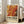 May include: Framed abstract art poster with swirling orange, red, yellow and black paint-like design displayed in bright living room corner next to beige upholstered chair. The vintage-style exhibition poster features bold "POLLOCK" text and dates "MAY 18, 2030 - AUGUST 14, 2030" below the chaotic, flowing artwork. Natural sunlight streams through white-framed windows onto light wooden floors.