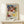 May include: Framed Paul Klee exhibition poster featuring abstract watercolor composition with overlapping geometric shapes in blues, reds, yellows, greens, and earth tones. The artwork shows varied textures including dotted and patterned sections. Poster displays museum information and artist name at bottom. Natural wood frame sits on light wooden shelf next to small white wrapped gift with twine and evergreen sprig against cream wall.