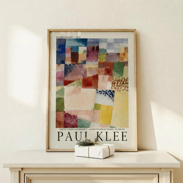 May include: Framed Paul Klee exhibition poster featuring abstract watercolor composition with overlapping geometric shapes in blues, reds, yellows, greens, and earth tones. The artwork shows varied textures including dotted and patterned sections. Poster displays museum information and artist name at bottom. Natural wood frame sits on light wooden shelf next to small white wrapped gift with twine and evergreen sprig against cream wall.