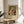 May include: Framed exhibition poster featuring Renoir's impressionist painting of a blonde woman in white dress holding a tabby cat, displayed on round wooden side table next to beige upholstered chair with white throw pillow. Soft natural lighting creates shadows on cream wall, with green plant leaves visible in corner.