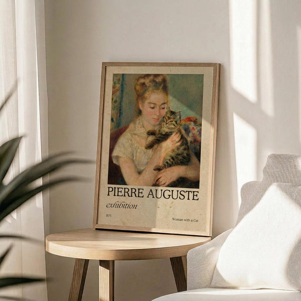 May include: Framed exhibition poster featuring Renoir's impressionist painting of a blonde woman in white dress holding a tabby cat, displayed on round wooden side table next to beige upholstered chair with white throw pillow. Soft natural lighting creates shadows on cream wall, with green plant leaves visible in corner.
