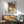 May include: Framed Paul Klee exhibition poster displaying colorful abstract geometric squares and rectangles in watercolor style, hanging above mid-century modern wooden credenza with white ceramic lamp. Sage green upholstered armchair with wooden frame sits beside small round side table. Bright natural sunlight creates geometric shadows on white wall, creating minimalist Scandinavian-inspired interior setting.