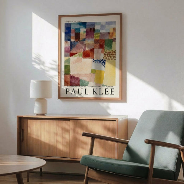 May include: Framed Paul Klee exhibition poster displaying colorful abstract geometric squares and rectangles in watercolor style, hanging above mid-century modern wooden credenza with white ceramic lamp. Sage green upholstered armchair with wooden frame sits beside small round side table. Bright natural sunlight creates geometric shadows on white wall, creating minimalist Scandinavian-inspired interior setting.