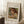 May include: Framed poster print of a Renoir painting showing a young child with reddish-brown hair reading a book, rendered in soft impressionist brushstrokes with warm earth tones. The beige matted frame leans against white wainscoting beside a wooden side table with coffee cup, on a textured cream rug next to a beige upholstered chair in natural lighting.