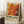 May include: Framed abstract art poster with vibrant orange, red, and yellow marbled paint swirls in Jackson Pollock style, leaning against white wall corner. Poster shows exhibition text "POLLOCK - EMBRACE THE CHAOTIC REALM OF CREATIVITY" with dates. Scene includes white coffee cup on wooden side table, cream textured rug, and beige throw blanket nearby. Natural window light creates soft shadows on the wall.