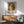 May include: Framed poster displaying a soft-toned impressionist painting of a blonde woman in white dress holding a striped tabby cat, hung above a wooden sideboard with white ceramic lamp in a modern living room with mint green armchair and natural lighting casting shadows on white walls.