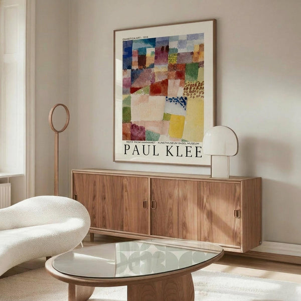 May include: Colorful Paul Klee abstract artwork poster displayed above wooden credenza in modern living room. The framed print features geometric squares and rectangles in vibrant blues, reds, yellows, and greens with varied textures and patterns. White curved chair and glass coffee table complete the minimalist Scandinavian-style interior with beige walls and natural lighting.