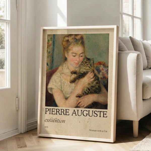 May include: Framed exhibition poster featuring Pierre Auguste Renoir's 1875 painting of a blonde woman in white clothing tenderly holding a striped tabby cat. The vintage-style poster has cream matting and light wood frame, leaning against white walls near large windows in a bright living room with beige sofa and pillows visible.