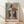 May include: Framed cubist-style portrait of a woman wearing a geometric hat with orange, black, and white triangular sections. The artwork features a pale face with dark hair, red lips, and is rendered in muted earth tones against a gray background. The poster is displayed in a light wood frame, leaning against a white wall on wooden flooring with natural sunlight casting diagonal shadows across the scene.