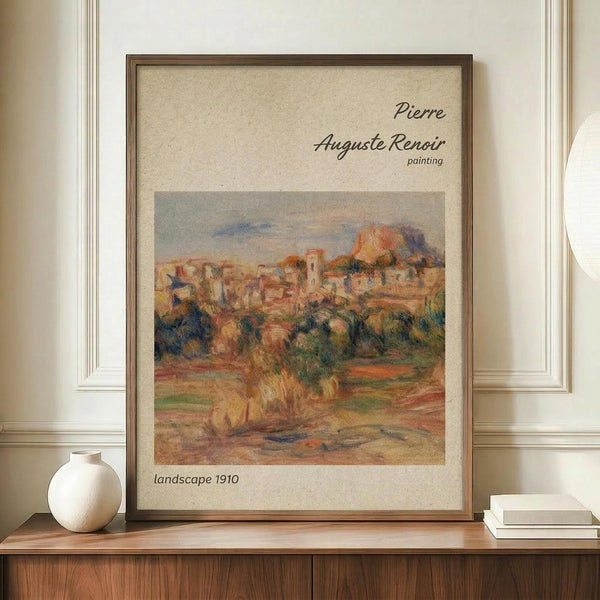 May include: Framed reproduction of Renoir's impressionist landscape painting showing a Mediterranean village with warm orange and terracotta buildings nestled among green foliage under a soft blue sky. The print features cream matting with black text identifying the artist and artwork date of 1910. Displayed on a wooden credenza alongside a white ceramic vase and stacked books against white wainscoting.