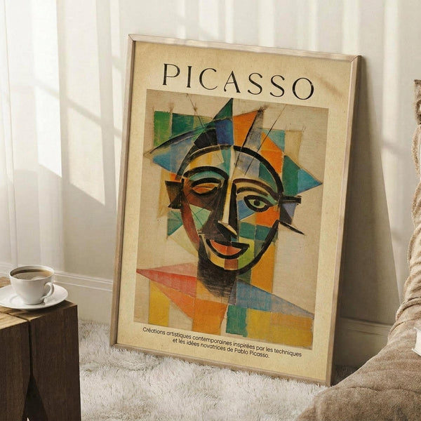 May include: Framed Picasso-style cubist portrait poster leaning against white wall corner on white textured rug. Abstract face features geometric shapes in vibrant blues, greens, oranges, and yellows on cream background. French text below artwork. Coffee cup on wooden block nearby, beige fabric visible at edge.