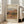 May include: Framed vintage-style art print featuring an impressionist landscape painting with warm earth tones showing Mediterranean hillside village with terracotta roofs and green vegetation. Print displays in light wood frame leaning against white wall beside beige upholstered armchair in bright, naturally-lit living room with wooden floors.