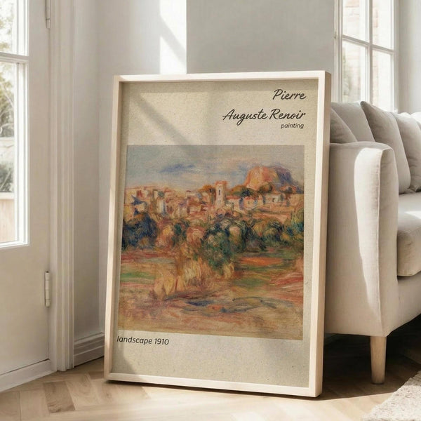 May include: Framed vintage-style art print featuring an impressionist landscape painting with warm earth tones showing Mediterranean hillside village with terracotta roofs and green vegetation. Print displays in light wood frame leaning against white wall beside beige upholstered armchair in bright, naturally-lit living room with wooden floors.