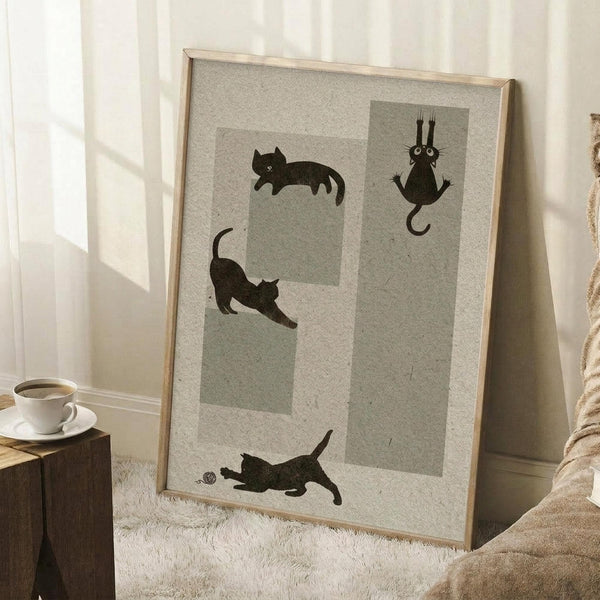 May include: Framed poster with four black cat silhouettes in different playful poses - sleeping, stretching, hanging, and playing with yarn - displayed on light wooden frame against white wall. Scene shows cozy interior with white coffee cup on wooden block side table and beige textured blanket, creating warm natural lighting atmosphere.