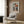 May include: Framed Picasso-style cubist portrait poster featuring woman with geometric orange and navy hat, displayed on wood paneled wall above beige upholstered armchair with patterned throw pillow. Modern brass floor lamp and dark side table with open book complete the neutral-toned reading nook scene.