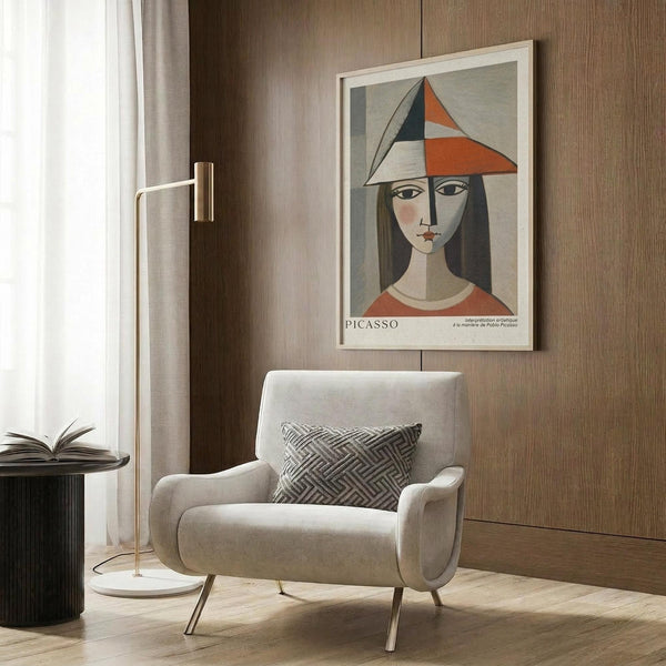 May include: Framed Picasso-style cubist portrait poster featuring woman with geometric orange and navy hat, displayed on wood paneled wall above beige upholstered armchair with patterned throw pillow. Modern brass floor lamp and dark side table with open book complete the neutral-toned reading nook scene.