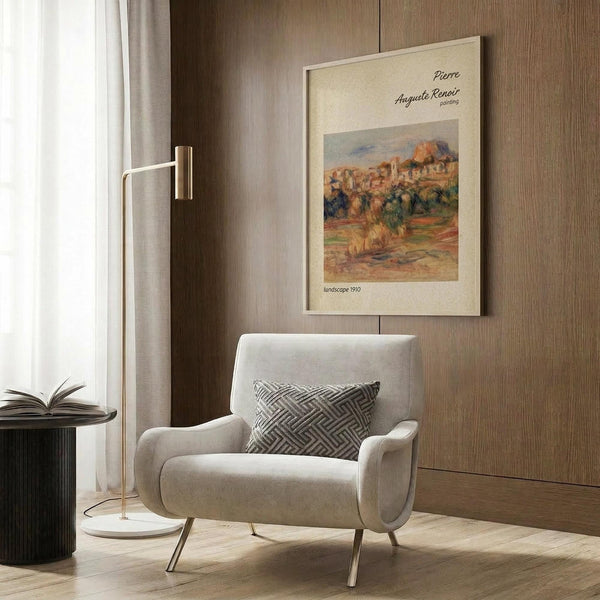 May include: Framed art print displayed on wood paneled wall above beige upholstered armchair with geometric patterned pillow. Print shows warm-toned landscape painting with buildings, trees, and rolling terrain in impressionist style. Modern reading nook setting with brass floor lamp, dark round side table with open book, and natural lighting from nearby window with curtains.