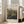 May include: Framed Renoir landscape painting print leaning against beige armchair in bright living room with white walls and windows. The artwork shows rolling hills with lush vegetation in impressionist style using greens, blues, and earth tones. Natural wood frame with cream matting displays the pastoral countryside scene with soft, dreamy brushwork typical of Renoir's plein air landscapes.
