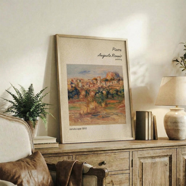 May include: Framed art print featuring a warm-toned landscape painting with rolling hills, trees, and Mediterranean-style buildings displayed on a rustic wooden sideboard. The beige matted print sits alongside potted plants, stacked books, ceramic vase, and table lamp in a cozy living room setting with neutral walls and upholstered furniture.