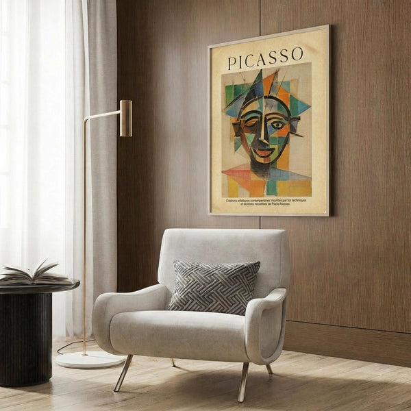 May include: Beige armchair with geometric patterned pillow positioned next to brass floor lamp and dark side table with open book. Framed Picasso-style cubist portrait poster hangs on wood-paneled wall, featuring abstract face in colorful geometric shapes with orange, blue, green, and yellow tones. Modern minimalist room setting with natural lighting from window and light wood floors.
