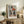 May include: Framed Picasso-style cubist portrait of woman wearing geometric angular hat in orange, navy, and cream colors, displayed on rustic wooden sideboard with fern plant, ceramic vase, stacked books, and beige upholstered chair in warm-toned living room setting