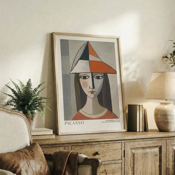 May include: Framed Picasso-style cubist portrait of woman wearing geometric angular hat in orange, navy, and cream colors, displayed on rustic wooden sideboard with fern plant, ceramic vase, stacked books, and beige upholstered chair in warm-toned living room setting