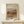 May include: Framed art print featuring an impressionist landscape painting with warm earth tones showing a hillside village with buildings and trees. The poster has a beige background with elegant script text and is displayed in a light wood frame on a white wooden surface next to a small white gift box with twine and greenery sprig against a cream wall.