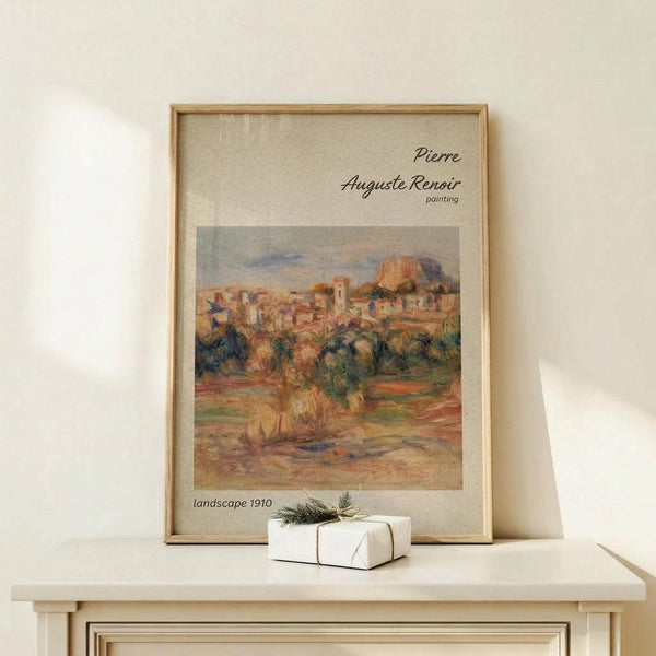 May include: Framed art print featuring an impressionist landscape painting with warm earth tones showing a hillside village with buildings and trees. The poster has a beige background with elegant script text and is displayed in a light wood frame on a white wooden surface next to a small white gift box with twine and greenery sprig against a cream wall.