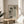 May include: Framed poster with minimalist illustration of four black cat silhouettes in playful poses on neutral beige background with geometric shapes, displayed on round wooden side table next to cream upholstered chair with plant shadows cast across white wall