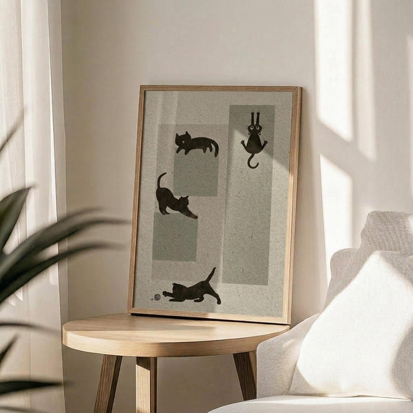 May include: Framed poster with minimalist illustration of four black cat silhouettes in playful poses on neutral beige background with geometric shapes, displayed on round wooden side table next to cream upholstered chair with plant shadows cast across white wall