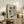 May include: Framed poster with minimalist illustration of three black cat silhouettes in different playful poses against geometric gray and beige background shapes, displayed on rustic wooden console table alongside fern plant, books, ceramic vase, and cream upholstered chair in neutral-toned living room setting.