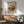 May include: Framed landscape poster print featuring warm earth tones and impressionist brushstrokes depicting buildings and foliage from 1910, displayed above wooden sideboard with white ceramic lamp and palm plant, next to mint green upholstered armchair with wooden arms in modern living room with natural sunlight streaming across white walls