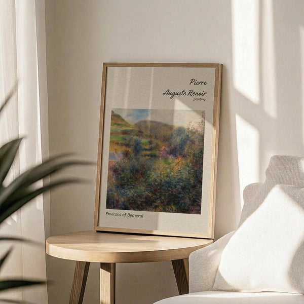May include: Framed art print featuring Pierre Auguste Renoir's impressionist landscape painting "Environs of Berneval" with rolling green hills, trees, and countryside scenery in soft blues and greens. Print displayed on light wood round side table next to beige upholstered chair and plant in bright, airy room with white walls and natural sunlight creating gentle shadows.