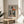 May include: Framed art print featuring a cubist-style portrait of a woman with geometric orange and black triangular hat, long dark hair, and coral-colored top against muted gray background. Print displays in thin gold frame on round wooden side table next to beige upholstered chair with decorative pillow. Soft natural lighting creates window shadows across white wall, with green plant leaves visible in corner.