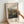 May include: Hands holding a light wood framed print against a white wall. The artwork shows an impressionist landscape painting with rolling hills, lush vegetation, and soft colors in greens, blues, and earth tones. Text above reads "Pierre Auguste Renoir painting" and below "Environs of Berneval" on cream colored matting.