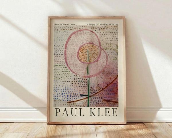 May include: Framed Paul Klee exhibition poster leaning against white wall on light wood floor. Abstract artwork features colorful dotted patterns surrounding a stylized flower with pink petals, orange center, and green stem. Soft natural lighting creates diagonal shadows across the scene. Light wooden frame complements the warm, minimalist interior setting.