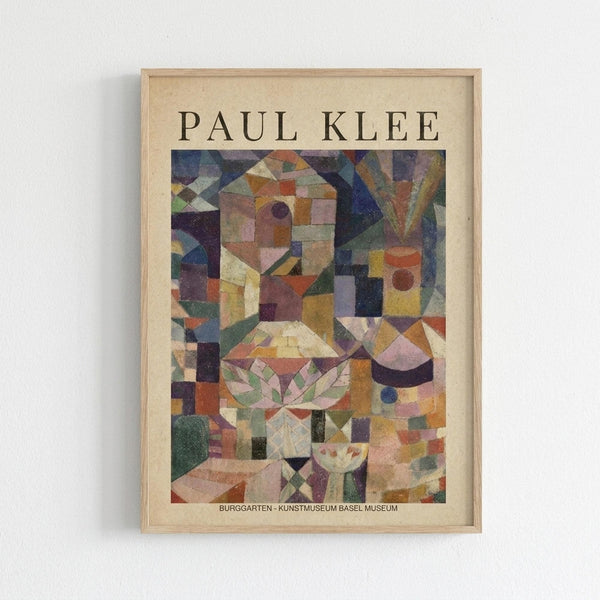 Framed art print featuring Paul Klee's abstract cubist painting with geometric shapes, colorful squares, and architectural forms in muted earth tones of orange, purple, green, and beige. The artwork shows fragmented buildings and structures in Klee's distinctive modernist style. Displayed in a light wood frame with cream matting against a white wall. Museum attribution text visible at bottom of print.