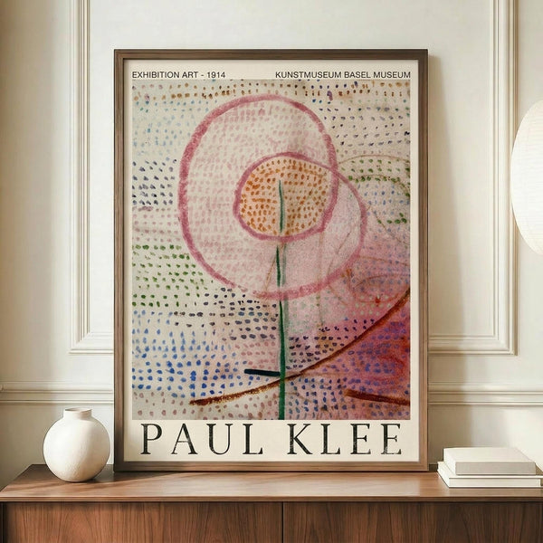 May include: Framed Paul Klee exhibition poster featuring abstract artwork with dotted patterns in blues, greens, and purples, pink circular forms, and orange center with green vertical line. Poster displayed on wooden credenza beside white ceramic vase and stacked books in neutral-toned room with white walls and crown molding.