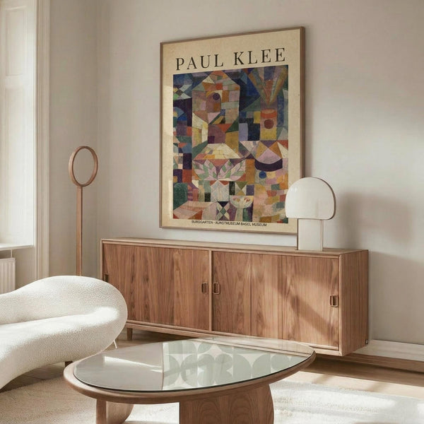 May include: Paul Klee abstract art poster in thin brown frame hanging above wooden sideboard in modern living room. Colorful geometric composition features blocks, triangles, and organic shapes in blues, oranges, greens, and earth tones. Room has cream walls, white sculptural chair, glass-top oval coffee table, and minimalist white table lamp.