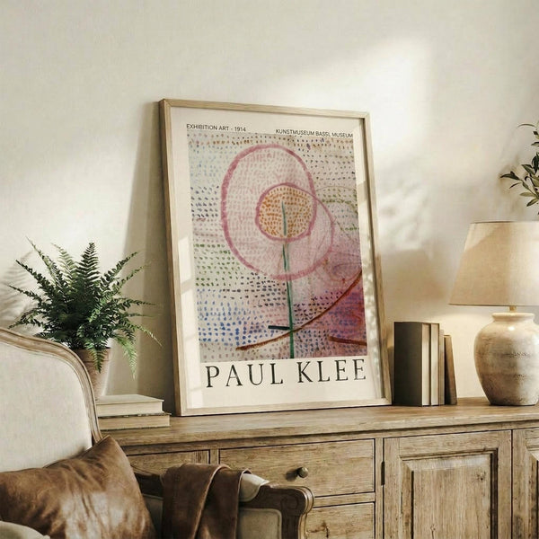 May include: Framed Paul Klee exhibition poster in light wood frame leaning against white wall on rustic wooden sideboard. Abstract artwork features pink and orange spiral flower design with dotted pattern background. Styled with fern plant, leather chair, stacked books, and ceramic vase in warm, minimalist living room setting.