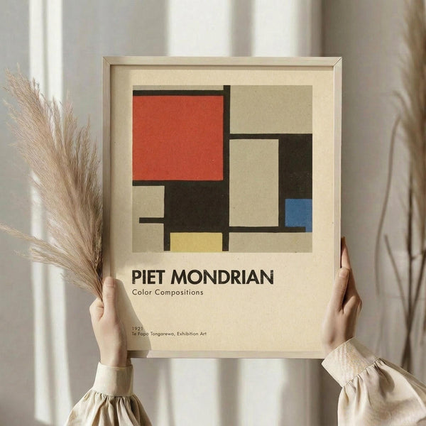 May include: Hands holding a framed geometric art print featuring rectangular blocks in red, blue, yellow, black, and cream colors separated by black lines, styled as a vintage exhibition poster. Person wears cream-colored clothing with gathered sleeves while holding dried pampas grass. Light wooden frame against neutral curtained background creates minimalist aesthetic.