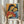 May include: Person in cream ruffled sleeves holding framed Picasso-style cubist artwork showing two embracing figures with geometric faces in warm yellows, oranges, reds and blues. Dried pampas grass visible on left. White curtained window background creates soft natural lighting. Silver frame contains vintage-style art poster with French text at bottom.