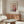 May include: Paul Klee exhibition poster in thin brown frame hanging above wooden credenza with sliding doors. Abstract floral artwork features pink and yellow circular blooms on dotted background with green stems. Room has cream walls, white curved chair, glass oval coffee table, and brass floor lamp. Natural lighting creates warm, minimalist Scandinavian interior styling.