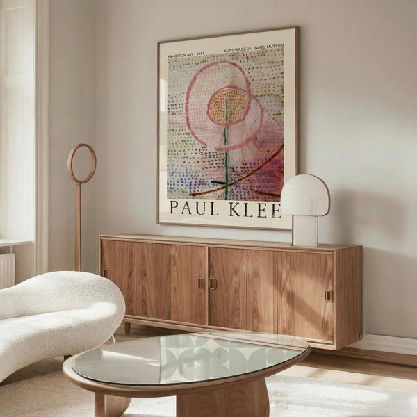 May include: Paul Klee exhibition poster in thin brown frame hanging above wooden credenza with sliding doors. Abstract floral artwork features pink and yellow circular blooms on dotted background with green stems. Room has cream walls, white curved chair, glass oval coffee table, and brass floor lamp. Natural lighting creates warm, minimalist Scandinavian interior styling.