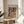 May include: Framed Paul Klee exhibition poster featuring abstract geometric composition with colorful overlapping shapes, houses, and botanical elements in vibrant blues, oranges, greens, and purples. Poster sits on round wooden side table next to cream upholstered chair with plant leaves visible in corner. Soft natural lighting creates shadows on white wall behind.