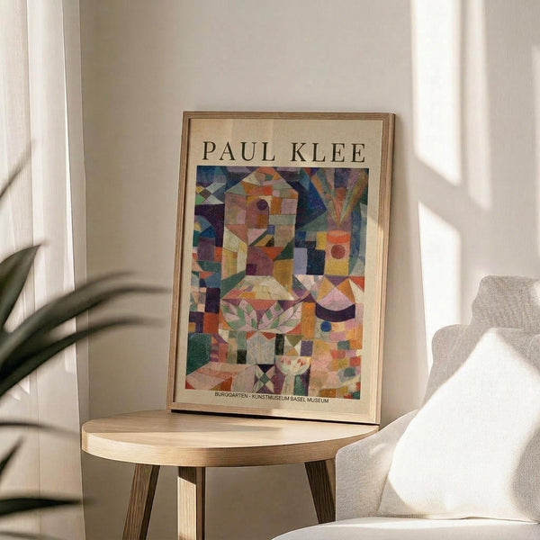 May include: Framed Paul Klee exhibition poster featuring abstract geometric composition with colorful overlapping shapes, houses, and botanical elements in vibrant blues, oranges, greens, and purples. Poster sits on round wooden side table next to cream upholstered chair with plant leaves visible in corner. Soft natural lighting creates shadows on white wall behind.